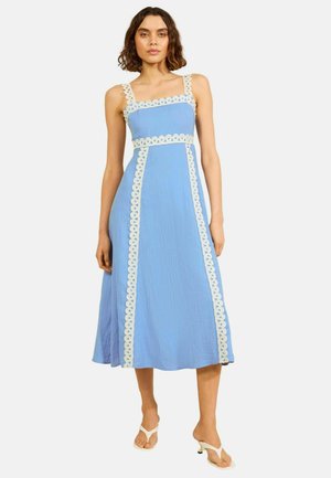 Nő, aki világoskék ujjatlan midi ruhát visel fehér hullámos szegéllyel, fehér magassarkú szandálokkal, világos háttér előtt.