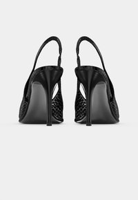 Sorte høyhælte slingback-sko med teksturert mønster på sidene, vist bakfra mot en ensfarget hvit bakgrunn.