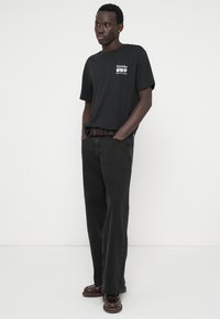 Μαύρο t-shirt με λευκό γραφικό στο στήθος, συνδυασμένο με σκούρο γκρι τζιν με ίσια γραμμή. Καφέ παπούτσια ολοκληρώνουν το σύνολο.