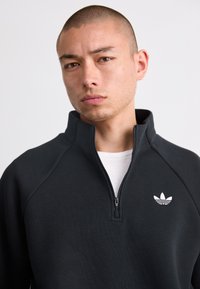 Sweat-shirt zippé noir avec manches raglan, fabriqué dans un tissu lisse ; arbore le logo Adidas blanc sur la zone gauche de la poitrine.