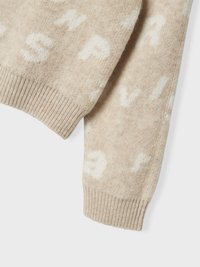 Beżowy wełniany sweter o miękkiej fakturze, z białymi literami w wzorze. Rękawy zakończone mankietami i luźny krój.