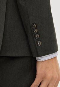 Zbliżenie na rękaw ciemnoszarego garnituru z czterema czarnymi guzikami na białym mankiecie koszuli oraz częściowo widoczną ręką.