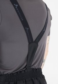 Zwarte stoffen bretels met een logo, bevestigd aan een grijze shirt. Het ontwerp heeft een gladde textuur en een verstelbare pasvorm met elastische banden.