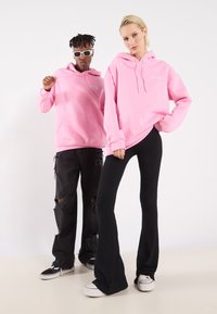 Due modelli indossano felpe rosa abbinati con tasca frontale e cappucci con coulisse. Il modello maschile abbina la sua con jeans neri strappati e sneakers.