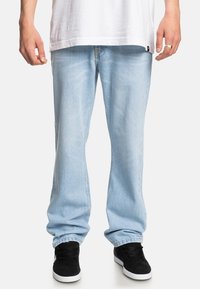 Hellblaue Denim-Jeans mit lockerem Schnitt, sichtbar genäht, mit leicht ausgewaschenem Finish und geraden Beinen. Getragen mit schwarzen Sneakern.
