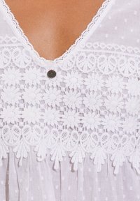 Vit blus med V-ringning, prydd med blommönsterbroderi, delikat spetskant och en dekorativ knapp med röd accent. Lätt, texturerad tyg.