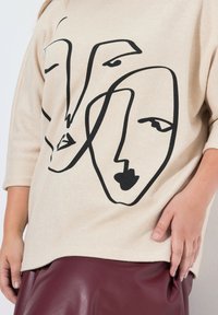 Beżowy sweter z trzema abstrakcyjnymi czarnymi twarzami nadrukowanymi z przodu. Posiada rękawy 3/4 i luźny, szeroki krój. Zestawiony z bordową skórzaną spódnicą.