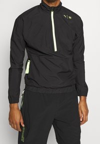 Man i svart Puma långärmad zip-up jacka och svarta shorts med neongröna dragkedje-accenter, stående framför en neutral bakgrund.