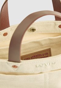Canvas tote bag met bruine leren handvatten, messing drukknoopsluiting en een Levi Strauss & Co. label aan de binnenzijde van de beige stoffen binnenkant.