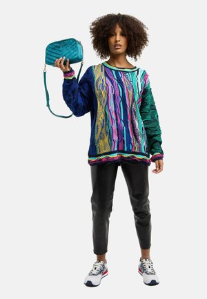 Frau mit lockigem Haar, die einen bunten, gemusterten Pullover, schwarze Lederhosen und weiße Sneakers trägt, hält eine türkisfarbene Handtasche mit erhobenem Arm.