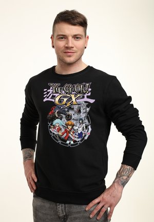 Mand med kort hår og tatoveringer iført sort Yu-Gi-Oh! GX-sweatshirt med tre animefigurer og en mekanisk drage.