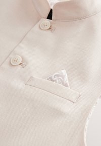 Vêtement beige clair avec un tissu texturé, comprenant un col montant, deux boutons blancs, et un mouchoir de poche blanc décoratif.