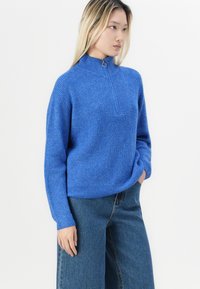 Maglione tricotato di un blu brillante, con colletto alto e chiusura a mezze zip. Presenta una texture a coste e una tasca sul petto. Abbinato a jeans a gamba larga.