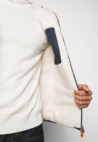 Marineblauwe jas met een pluizige beige voering. Bevat een ritszak aan de binnenkant van de linkerzijde en een oranje trekker aan de rits.