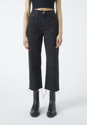 Osoba nosząca czarne, krótkie jeansy z wysokim stanem i postrzępionymi krawędziami, czarne botki z kwadratowym noskiem oraz czarny krótki top, stojąca na białym tle.