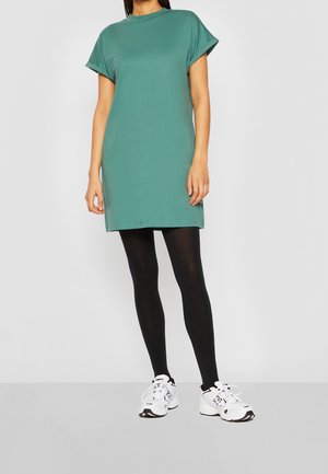 Vrouw die een turquoise jurkje met korte mouwen draagt, zwarte panty's en witte sneakers, staand tegen een effen lichtgrijze achtergrond.