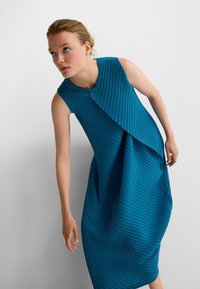 Ein türkisfarbenes, plisseeartiges Kleid mit ärmellosem Design, das eine asymmetrische Überlage und eine Vorderknopfleiste aufweist, aus strukturiertem Stoff gefertigt.