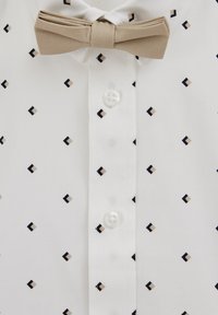 Beige rusetti valkoisella paidalla, jossa pieniä mustia ja beigejä timantinmuotoisia kuvioita napinaukossa.