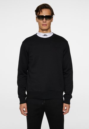 Sort sweatshirt med ribbede manchetter og kant, med et diskret logo på brystet. Båret med et hvidt underlag med høj krave og solbriller.