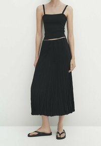 Nainen, jolla on päällään musta ilmanhiha crop-top, pitkä musta laskoshame ja mustat litteät sandaalit, seisoo tasaista valkoista taustaa vasten.