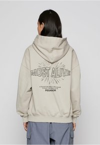 Beež kapuutsiga dressipluus graafilise kujundusega "MOST ALIVE" ja tekstiga tagaküljel, mis on lõdvestunud lõikega ning soonikuga varrukasäärtega.