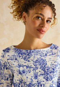 Weißes Oberteil mit einem blauen Landschaftsmuster mit Bäumen und Gebäuden. Rundhalsausschnitt, strukturiertes Material und figurbetontes Design.