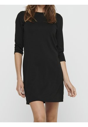 Femme portant une robe noire simple longueur genou avec des manches trois-quarts sur un fond clair uni.