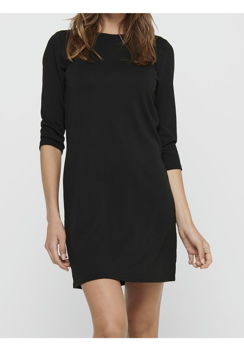 Femme portant une robe noire simple longueur genou avec des manches trois-quarts sur un fond clair uni.