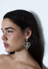 Giovane donna con capelli scuri indossa grandi orecchini di ferro a forma di cuore argentati, rivolta a sinistra su uno sfondo bianco.
