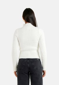 Weißer gerippter Strickpullover mit hohem Kragen und langen Ärmeln, der eine weiche Textur und eine etwas verkürzte Passform aufweist, kombiniert mit dunklen Jeans.