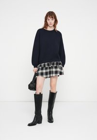 Sweat-shirt surdimensionné bleu marine, jupe à carreaux en couches, bottes noires montantes avec un talon carré et un petit sac banane noir.