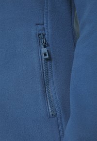 Granatowa kurtka polarowa z bocznym zamkiem błyskawicznym. Gładka tekstura z przeszywanymi szwami i eleganckim krojem. Widoczne trwałe elementy zamka.