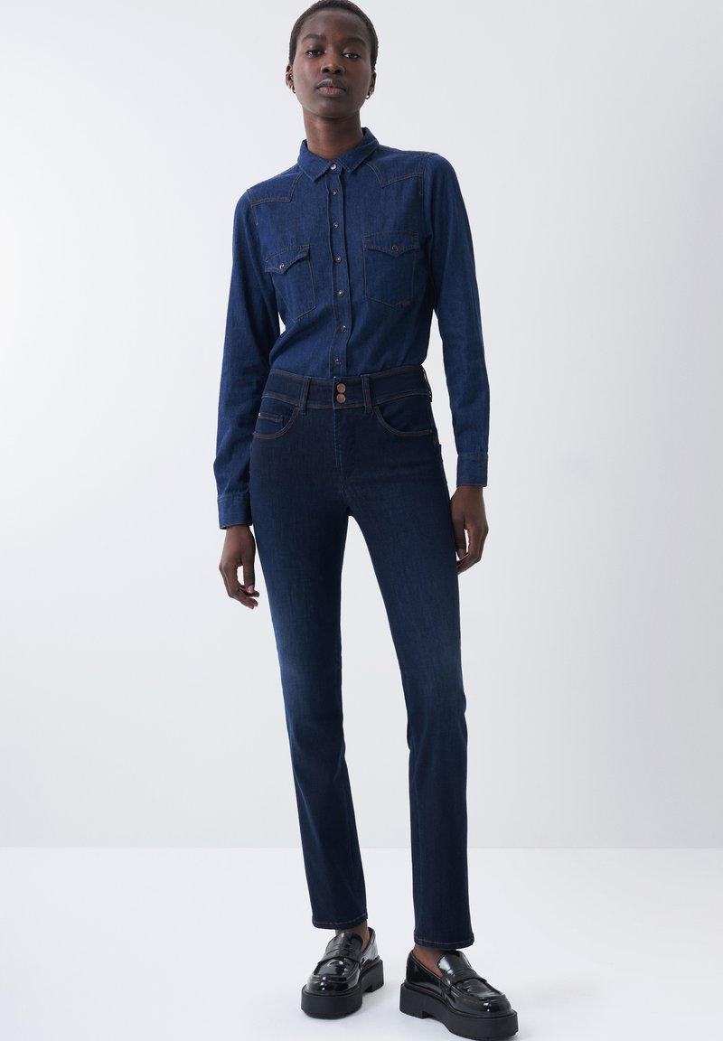 Camisa de mezclilla oscura con tapeta de botones y dos bolsillos en el pecho, combinada con jeans oscuros de corte ajustado y zapatos de plataforma negros, sobre un modelo contra un fondo simple.