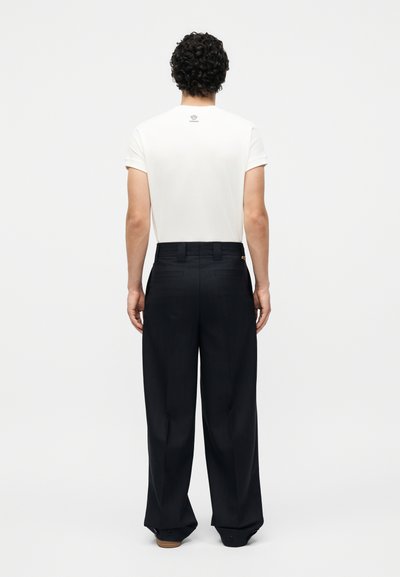 Άνδρας με σγουρά μαλλιά που φοράει λευκό κοντομάνικο μπλουζάκι και φαρδιά μαύρη παντελόνια, στέκεται με την πλάτη σε απλό φόντο.