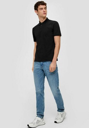 Schwarzes Polohemd mit einer Knopfleiste mit drei Knöpfen, kombiniert mit hellblauen Jeans und weißen Turnschuhen. Der Stoff wirkt glatt und hat einen figurbetonten Schnitt.