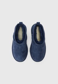 Pantuflas de ante azul marino con suave forro de piel, diseño deslizante, punta redondeada y tacón pequeño. Presentan un acento de costura a lo largo del borde.