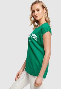 Grøn t-shirt i bomuld med korte ærmer, med hvid "NEW YORK" tryk på tværs af brystet; afslappet pasform og let buet nederste kant.
