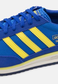 Blauwe en gele sportschoen van mesh-stof, met drie diagonale strepen, textuuraccenten en een rubberen buitenzool voor grip.