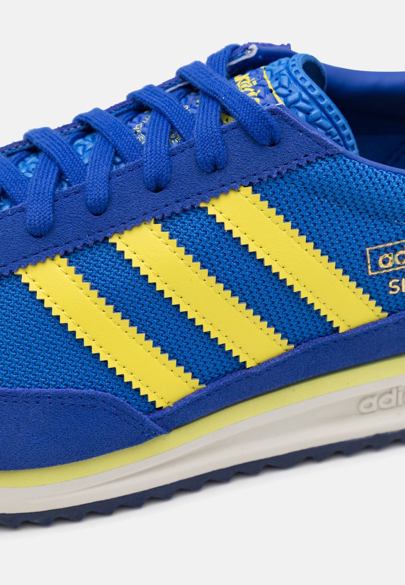 Blauer und gelber Sportschuh aus Netzgewebe, mit drei diagonalen Streifen, strukturierten Akzenten und einer Gummisohle für besseren Halt.