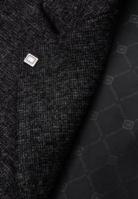 Tissu en tweed gris avec une finition texturée, présentant une épingle carrée argentée et une doublure noire lisse arborant un motif de logo discret.