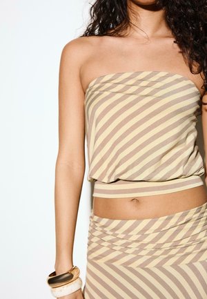 Kvinde iført stropløs beige og brun diagonalt striped top og matchende nederdel, med tre chunky armbånd på venstre håndled.