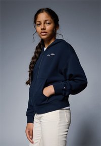 Marineblå zip-hettejakke laget av bomull-blandingsstoff, med en frontlomme og brodert logo på brystet; kombinert med kremfargede bukser.