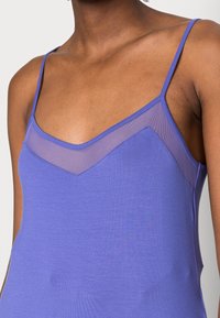 Närbild av en person som bär en ärmlös lila topp med tunna band och en genomskinlig panel över övre delen av bröstet.