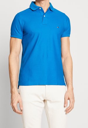 Homme portant un polo bleu vif à manches courtes et un pantalon blanc, debout devant un fond clair uni.