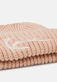 Berretto lavorato a maglia di un morbido colore pesca con texture a coste, dotato di un logo ricamato bianco. Il design con risvolto conferisce calore e stile.