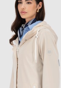 Giacca impermeabile beige con cappuccio, chiusura con zip, cordoncini e dettaglio con logo sul maniche, abbinata a una camicia a righe blu e bianche sotto.