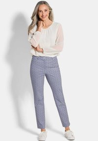 Witte blouse met geplooide halslijn, blauwe broek met een geometrisch patroon en witte sneakers tegen een effen achtergrond.