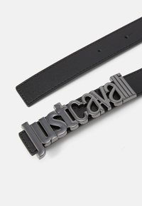 Svart belte med en jevn tekstur, med en fremtredende metallgrå logo "Just Cavalli" som spenne. Minimalistisk og moderne design.