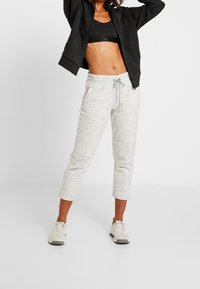 Pantaloni jogger grigio chiaro, taglio corto, in un tessuto texturizzato con vita regolabile e tasche laterali, abbinati a un reggiseno sportivo nero e sneakers grigie.