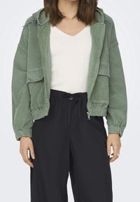 Veste bomber en velours côtelé vert avec capuche, présentant des poignets côtelés, une fermeture éclair à l'avant et deux poches avant. Portée par-dessus un haut blanc à côtes.